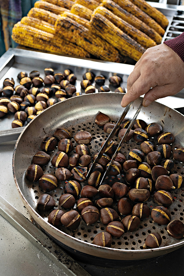 Ένας καστανάς γυρνάει με την τσιμπίδα τα κάστανα που ψήνονται στη φουφού. Δίπλα τους έχει μια ντάνα με ψητά καλαμπόκια.