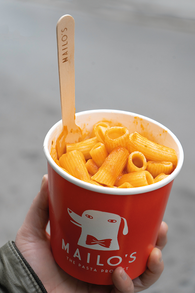 Ένα χέρι κρατάει μια χάρτινη συσκευασία με φρέσκα ζυμαρικά με ντομάτα και βασιλικό από τα Mailo's.